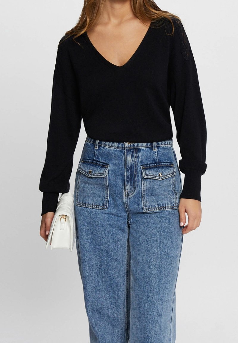 Mujer con un suéter negro de cuello en V y jeans de tiro alto azul con bolsillos delanteros, sosteniendo un bolso clutch blanco en una mano.
