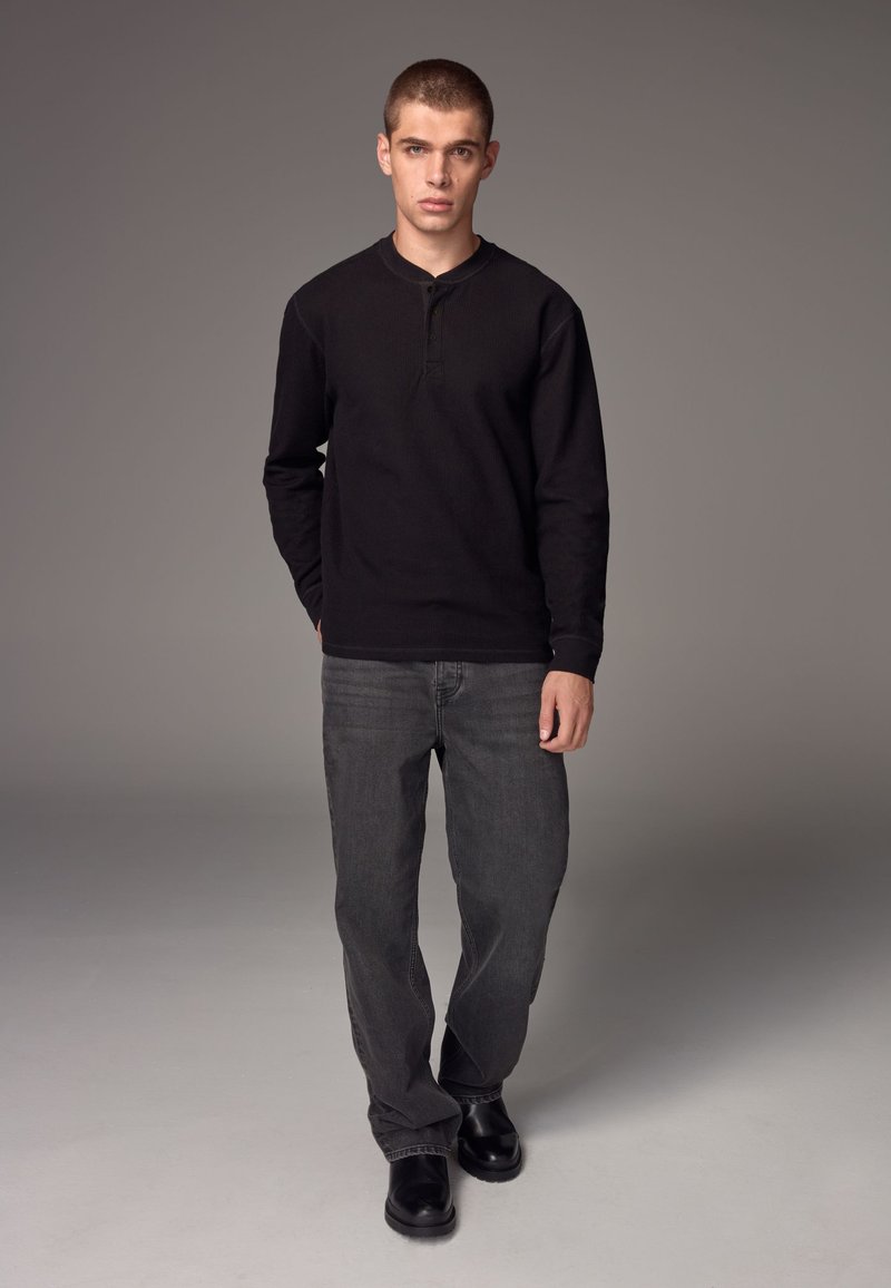 Schwarzes, geripptes Langarm-Henley-Shirt, graue Jeans und schwarze Schuhe. Das Modell steht aufrecht vor einem schlichten grauen Hintergrund.