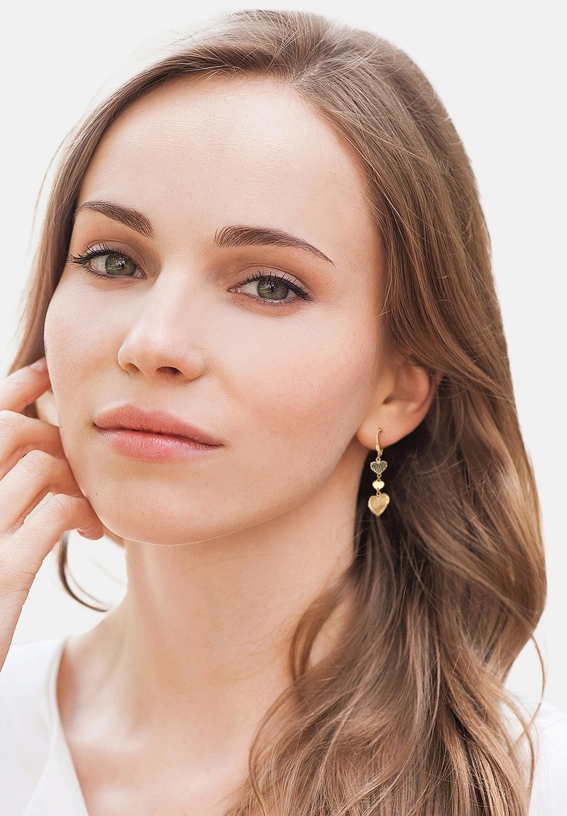 Guldbelagte øreringe med hjerteformer, der har et lagdelt design. Modellen har langt, bølget hår og en neutral, minimalistisk påklædning.