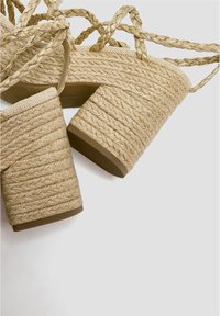 Talons blocs tressés beiges avec un dessus texturé, dotés d'une large bride et d'une semelle épaisse, complétés par des accents tressés entrelacés.