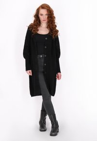 Schwarzer gestrickter langer Cardigan mit Knopfverschluss, gerippten Bündchen und offenen Struktur-Details an den Ärmeln, kombiniert mit grauen Jeans und schwarzen Stiefeln.