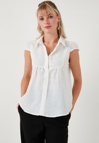 Bílá košile s krátkým rukávem, zapínání na knoflíky, strukturovaný design a měkký materiál. V kombinaci s černými kalhotami.