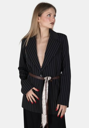 Donna che indossa un blazer nero a righe sottili con cintura marrone e sciarpa fantasia annodata, guarda di lato con smalto rosso e orecchini a cerchio dorati.