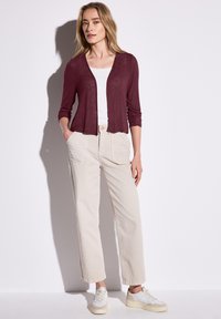 Vinröd lätt cardigan över en vit topp, tillsammans med lösa beige byxor och vita sneakers. Tyget verkar mjukt med en avslappnad passform.