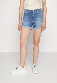 Shorts en denim de couleur bleu clair avec un ourlet roulé, dotés de cinq poches et d'une fermeture à bouton à l'avant, accompagnés de baskets blanches.