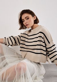 Pull en maille beige avec des rayures noires, coupe décontractée et col côtelé. porté sur une jupe en tulle blanc transparent, créant une texture superposée.