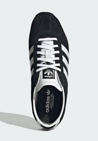 Melnas zamšādas kedas ar baltām ādas svītrām, baltām auklām un melnu mēli, kurā redzams Adidas logo; gumijas zole.