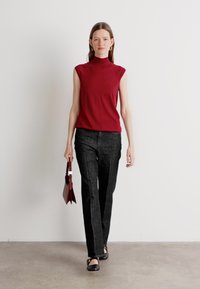 Top rosso senza maniche, pantaloni neri e scarpe nere. La modella porta una piccola borsa a mano rossa. Tessuto morbido e design aderente con colletto alto.