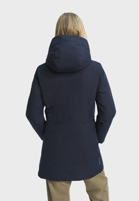 Osoba u tamnoplavoj jakni s kapuljačom i bež hlačama, stoji s rukama u džepovima, okrenuta leđima prema jednostavnoj svijetloj pozadini.