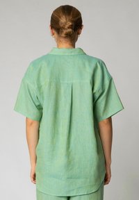Camicia verde chiaro a maniche corte con colletto, realizzata in un tessuto testurizzato e dotata di una piega centrale sul retro. Vestibilità morbida e rilassata con spalle scese.