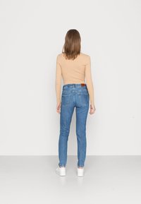 Top de mangas longas em bege claro com textura canelada, combinado com jeans de cintura alta azul médio. Os jeans apresentam bolsos traseiros e um patch em couro castanho. Ténis brancos.