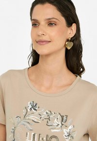 T-shirt beige avec un design floral en sequins, décolleté rond, porté avec des boucles d'oreilles en forme de cœur dorées. Tissu lisse, manches courtes, coupe décontractée.