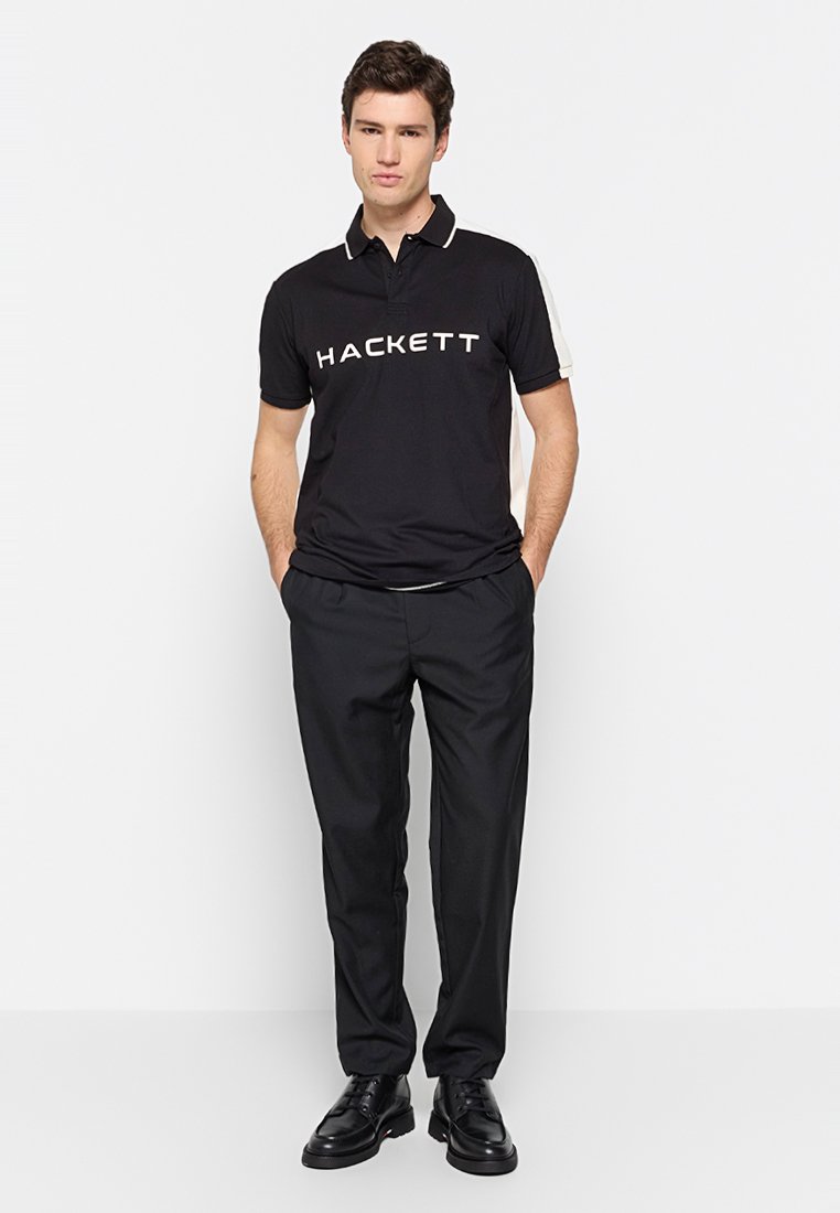 Schwarzes Polohemd mit weißem "HACKETT"-Logo, kurzes Oberteil mit kontrastierenden weißen Akzenten, kombiniert mit schwarzen Hosen und schwarzen Schuhen.