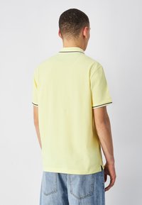 Koszulka polo w jasnożółtym kolorze z bawełny, z czarnymi wykończeniami na kołnierzyku i rękawach. Krótkie rękawy, luźny krój i rozporki po bokach u dołu.