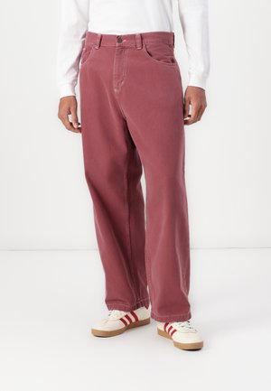 Losse, bordeauxrode katoenbroek met knoopsluiting, riemlussen en zijzakken, gecombineerd met witte sneakers met rode accenten.