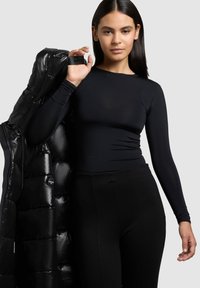 Sort langærmet top med tætsiddende design, sammen med sorte høj taljede bukser og en skinnende sort dunjakke hængende over den ene skulder.