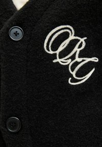 Juodas vilnonis kardiganas su tekstūruotu paviršiumi, juodais mygtukais ir baltais siuvinėtais inicialais "ORG" priekyje.