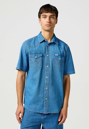 Kurze Ärmel Denim-Hemd in Hellblau. Mit zwei Brusttaschen mit Zickzackstickerei, einem Kragen und Knopfverschluss. Glatte Textur.