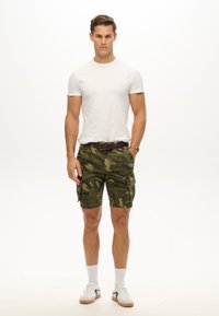 Camo cargo shorts i grøn med lommer, parret med en hvid t-shirt og hvide ankelsokker. Skoene er hvide med sorte detaljer.