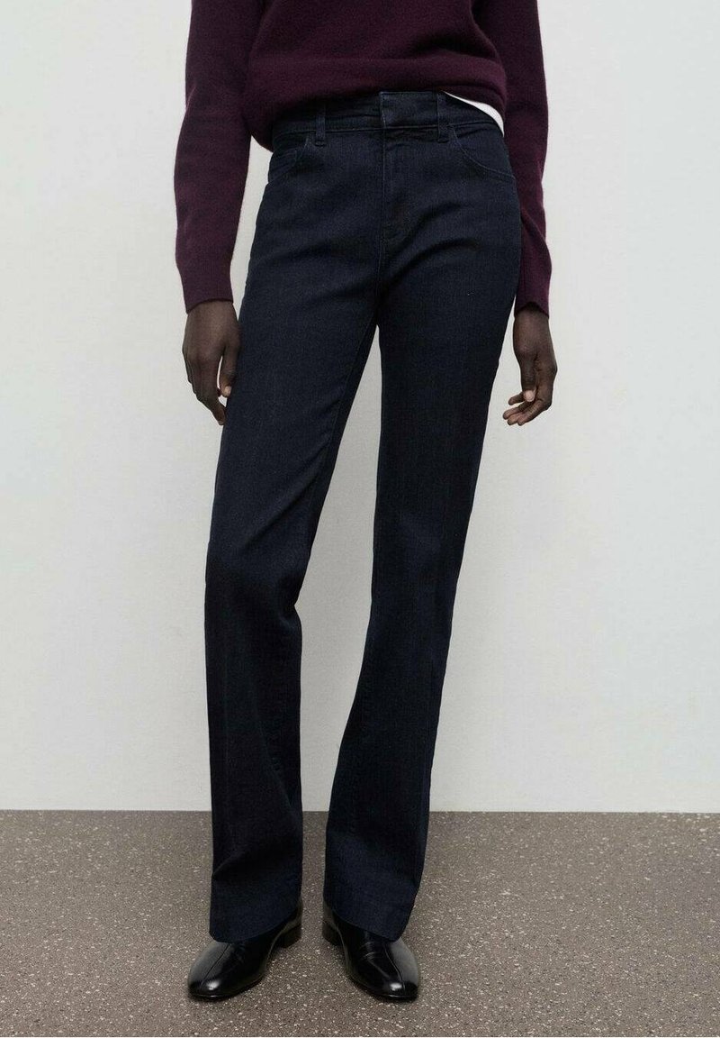 Personne portant un jean droit bleu foncé, des chaussures noires et un pull bordeaux foncé, debout contre un mur uni.