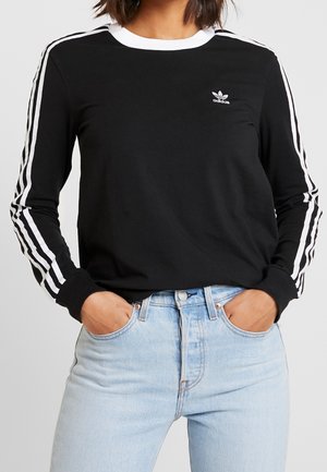 Personne portant un haut à manches longues noir Adidas avec des bandes blanches sur les manches et le logo sur la poitrine, associé à un jean bleu clair.