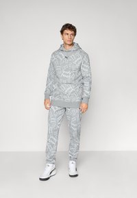 Mand iført grå hættetrøje og joggingbukser med hvidt zigzag-mønster samt hvide sneakers, der står foran en ensfarvet lys baggrund.