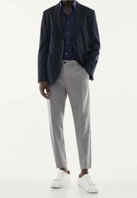 Pantaloni grigi su misura, giacca scura e camicia blu navy. Sneakers bianche. La giacca ha una chiusura con bottoni e tasche, con una texture liscia in tutto.