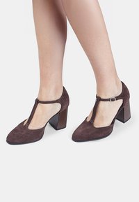 Zapatos de tacón de ante marrón con un diseño de tira en forma de T, lados abiertos y un tacón bloque apilado. Textura suave con una punta en forma de punta.