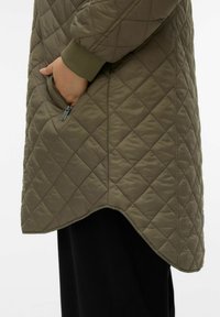 Manteau matelassé vert olive avec couture diagonale, doté d'une poche à main zippée et d'un ourlet arrondi. Extérieur doux et texturé avec poignets côtelés.
