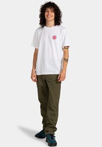 Weisses Baumwoll-T-Shirt mit einem runden grafischen Logo in Rot und Blau auf der Brust, kombiniert mit olivgrünen Hosen und schwarzen Schuhen mit blauen Akzenten.