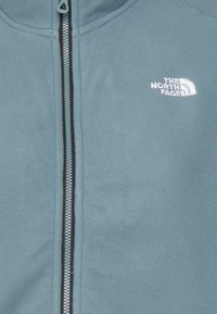 Jasnoniebieska polarowa kurtka z pełnym zamkiem błyskawicznym, czarnymi detalami zamka i białym logo na górnej lewej części klatki piersiowej. Miękka faktura.