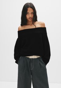 Czarny sweter typu off-the-shoulder z prążkowanym dekoltem, zestawiony z luźnymi ciemnymi dżinsami i jasnym paskiem z metalową klamrą.