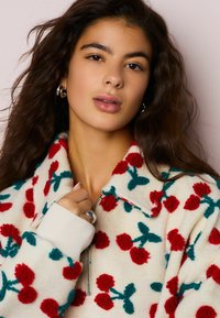 Jopica z zadrgo v smetanasti barvi z rdečimi cvetličnimi vzorci, zelenimi poudarki in rebrastimi belimi manšetami. Mehka tekstura, sproščen kroj in pogled na zgornjo polovico.