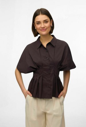 Kobieta o krótkich brązowych włosach, ubrana w czarną, zapinaną na guziki bluzkę z bufkami i kremowe spodnie o szerokich nogawkach, z rękami w kieszeniach, uśmiechająca się.