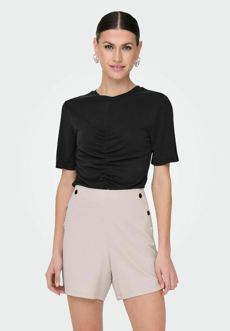 Top negro de manga corta con detalles fruncidos en la parte frontal, combinado con pantalones cortos de talle alto en color rosa claro que presentan acentos de botones dorados en los laterales.