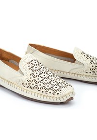 Chaussures slip-on en cuir beige avec des découpes florales délicates, des coutures le long de la semelle et une texture lisse.