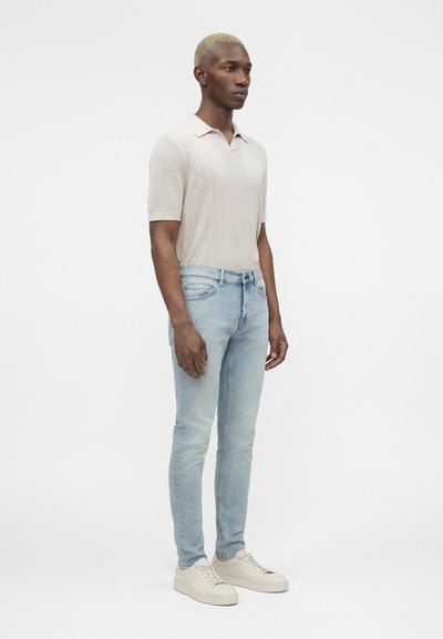 Άνδρας που στέκεται, φοράει ανοιχτό μπεζ πλεκτό πόλο με κοντά μανίκια, στενά ανοιχτογάλαζα τζιν και εκρού αθλητικά παπούτσια μπροστά σε απλό φόντο.