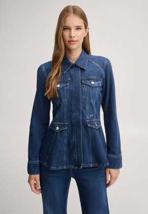 Frau mit langen, glatten Haaren trägt eine taillierte dunkelblaue Jeansjacke mit vier Fronttaschen und steht vor einem schlichten, hellen Hintergrund.
