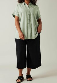 Hellgrünes Hemd mit kurzen umgeschlagenen Ärmeln und einer Brusttasche, kombiniert mit schwarzen, weit geschnittenen, knöchellangen Hosen und schwarzen Sandalen.