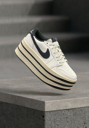 Hvid og sort Nike sneaker med tyk stribet platformsål, svævende over en betonhylde mod en sløret mørk baggrund.