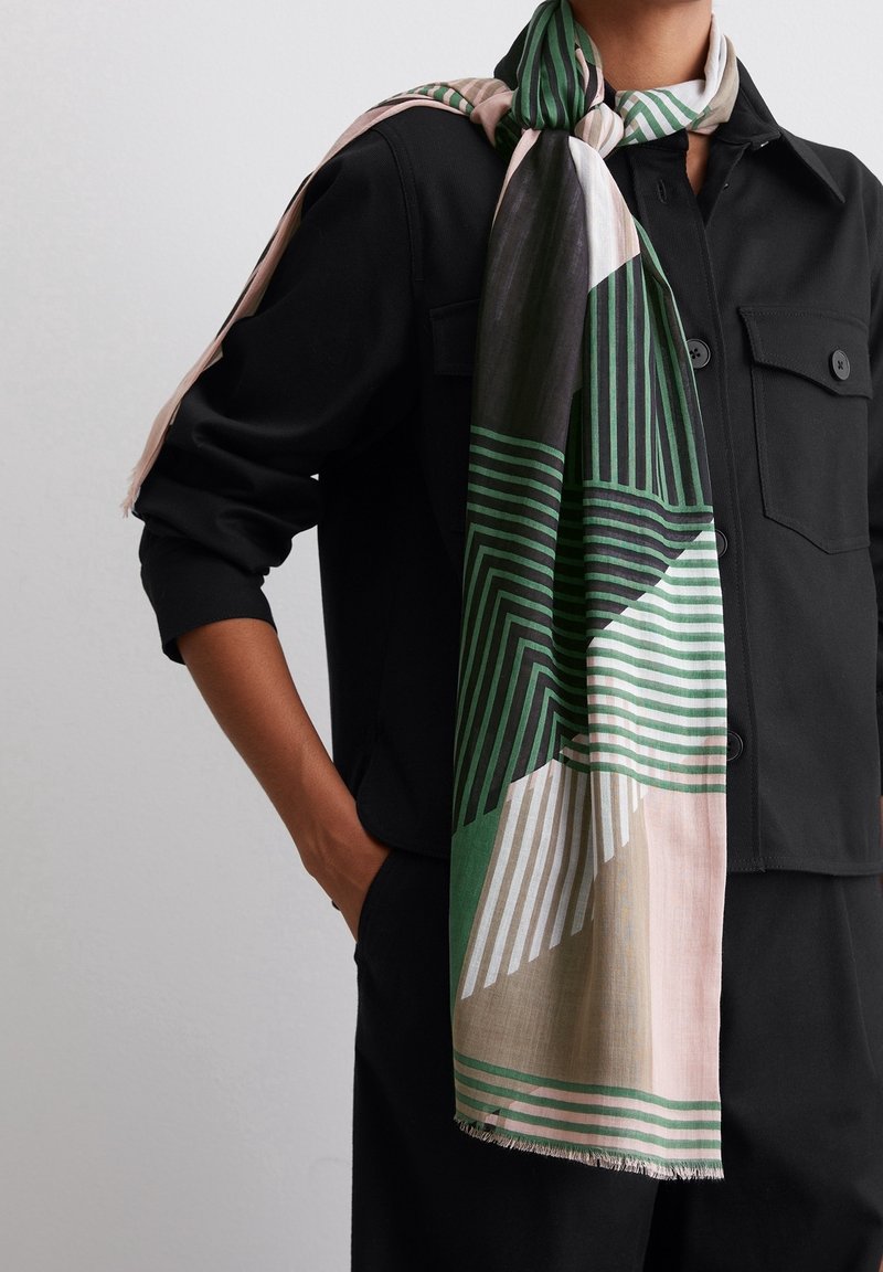 Personne portant une veste noire avec une grande écharpe à rayures géométriques vertes, noires, beiges et roses drapée sur l'épaule et autour du cou.