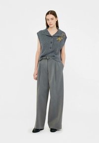 Graues, gestricktes ärmelloses Oberteil mit Kragen und drei Knöpfen; kombiniert mit locker sitzenden grauen Hosen und einer dekorativen Gürtelschnalle.