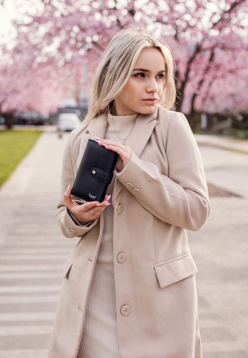 Black leather wallet with snap closure, held by a person in a beige coat, against a backdrop of pink flowering trees. Smooth texture, compact design.