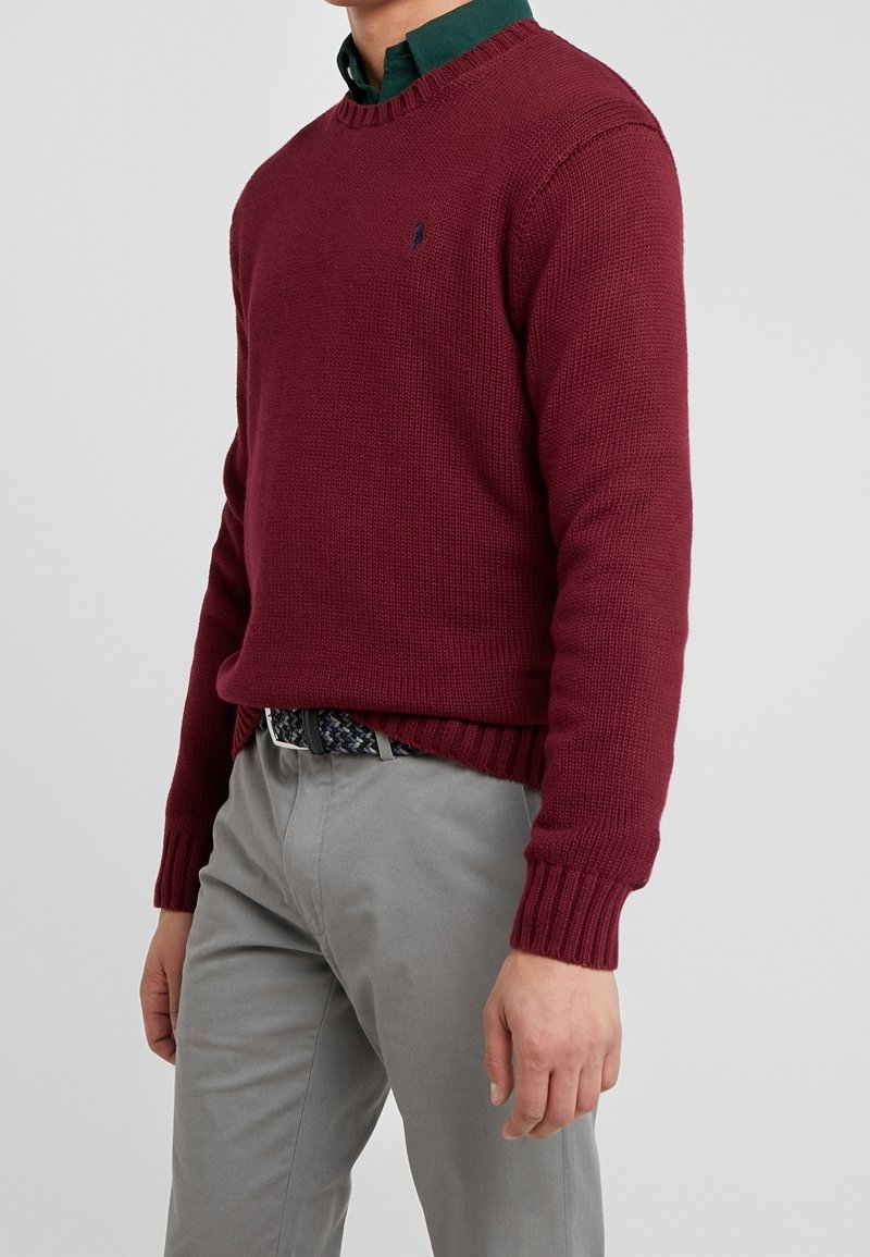 Homme portant un pull en maille bordeaux par-dessus une chemise verte à col, pantalon gris et ceinture tressée, debout devant un fond clair uni.