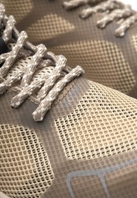 Beige mesh idrottsskor med texturerade detaljer, platta skosnören och en flexibel sula. Designen inkluderar mönstrade överlägg för andningsförmåga.