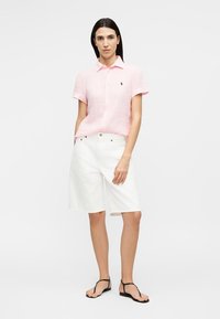 Femeie purtând o cămașă roz pal cu mâneci scurte și nasturi, pantaloni scurți albi până la genunchi și sandale negre, stând în picioare pe un fundal simplu.