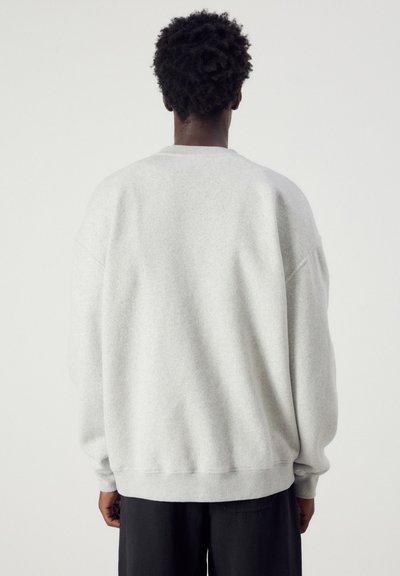 Personne aux cheveux courts et bouclés portant un sweat-shirt à manches longues gris clair uni et un pantalon foncé, vue de dos sur un fond uni.