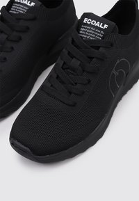 Zapatillas negras con parte superior de malla texturizada, puntera redonda y cordones planos. Cuenta con un logo en el lateral y un cuello acolchado para mayor comodidad.
