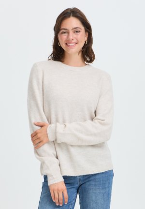 Jeune femme souriante, portant un pull beige clair et un jean bleu, debout devant un fond blanc uni.