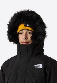 Μαύρο μπουφάν πάπλωμα με κουκούλα επενδεδυμένη με συνθετική γούνα, με μπροστινό φερμουάρ και λογότυπο The North Face. Φορεμένο με φωτεινό κίτρινο πλεκτό σκουφάκι.
